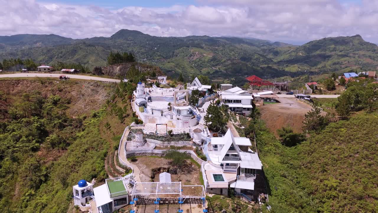 Wide aerial panorama of a scenic mountain resort with white Greek-inspired architecture. A unique tourist destination perched on a green hillside overlooking a lush valley and winding road