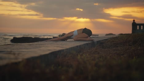 woman performs child pose on shore of bali with dramatic sunset in background