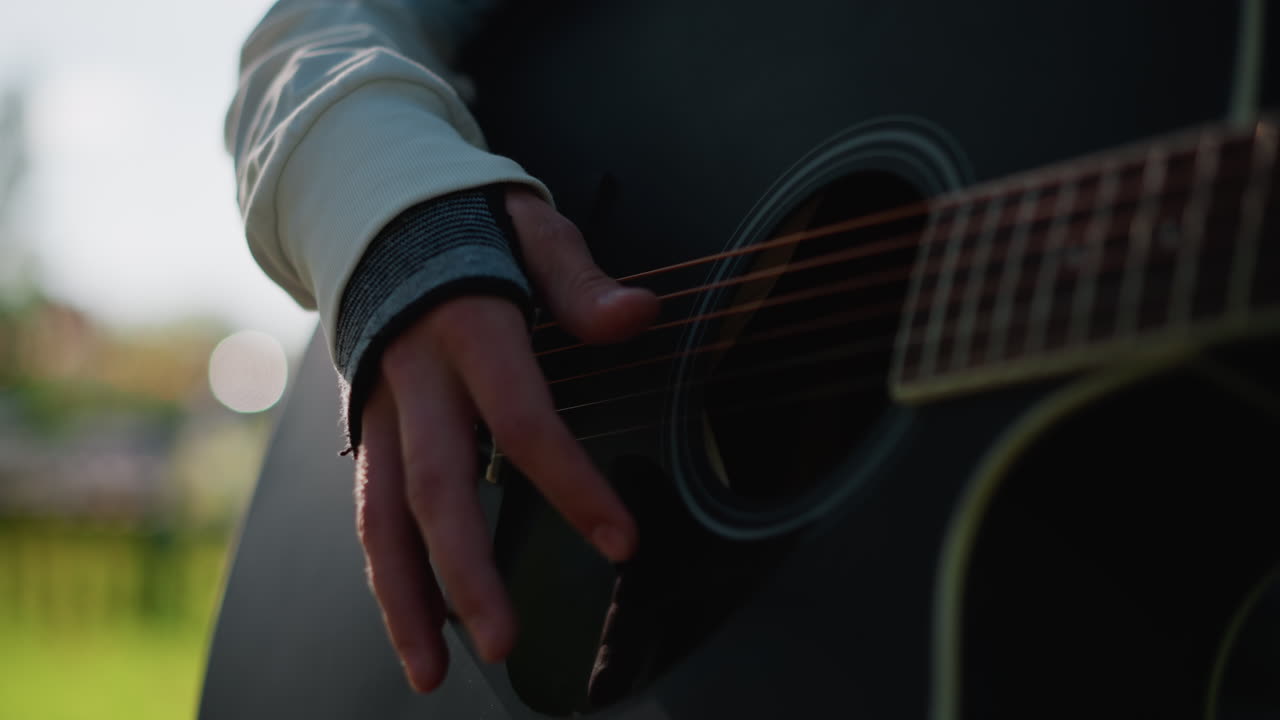 artista di strada che suona la chitarra acustica all'aperto in un parco, figura sfocata in primo piano con chitarra, staccionata rossa e area giochi sullo sfondo, erba verde brillante alla luce del sole, atmosfera spontanea di spettacolo di strada, allegro