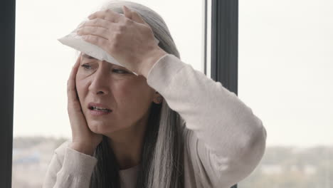 sick woman having headache and holding an ice pack to her head while sitting by the window at home 1