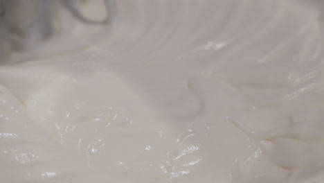 close up macro view of metal mixer stirring and blending thick, white icing in large bowl in slow motion