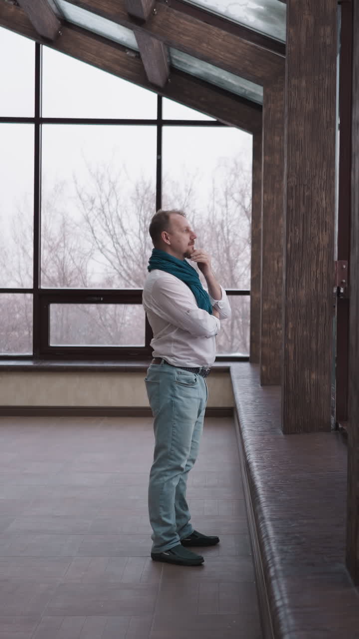 hombre mira el paisaje de invierno fuera de la ventana panorámica. pensativo caballero maduro se encuentra solo junto a la pared de vidrio en la casa residencial. tranquila soledad