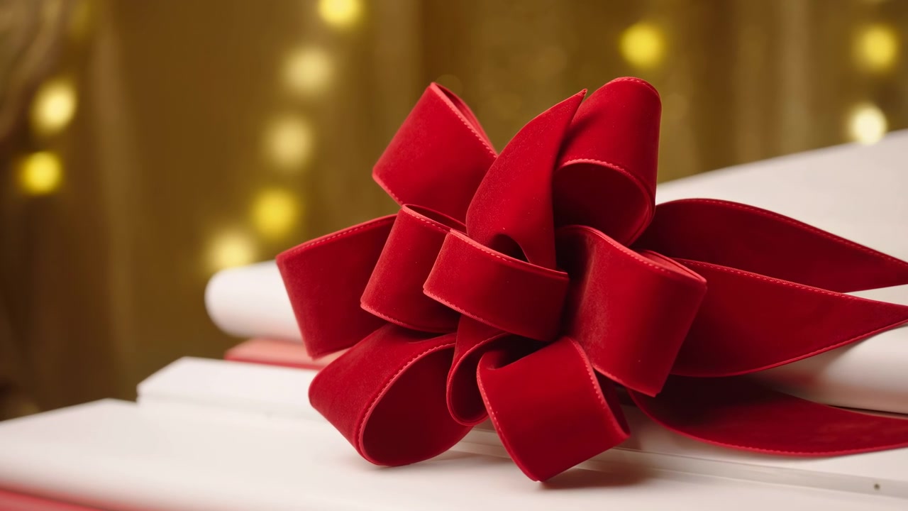 Close low-angle motion shot of large red velvet bow positioned at right foreground on stacked white gift boxes, showing multiple loops and pointed tails