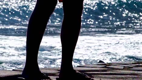 legs of a man wearing flip-flops at the sea side, big waves in background