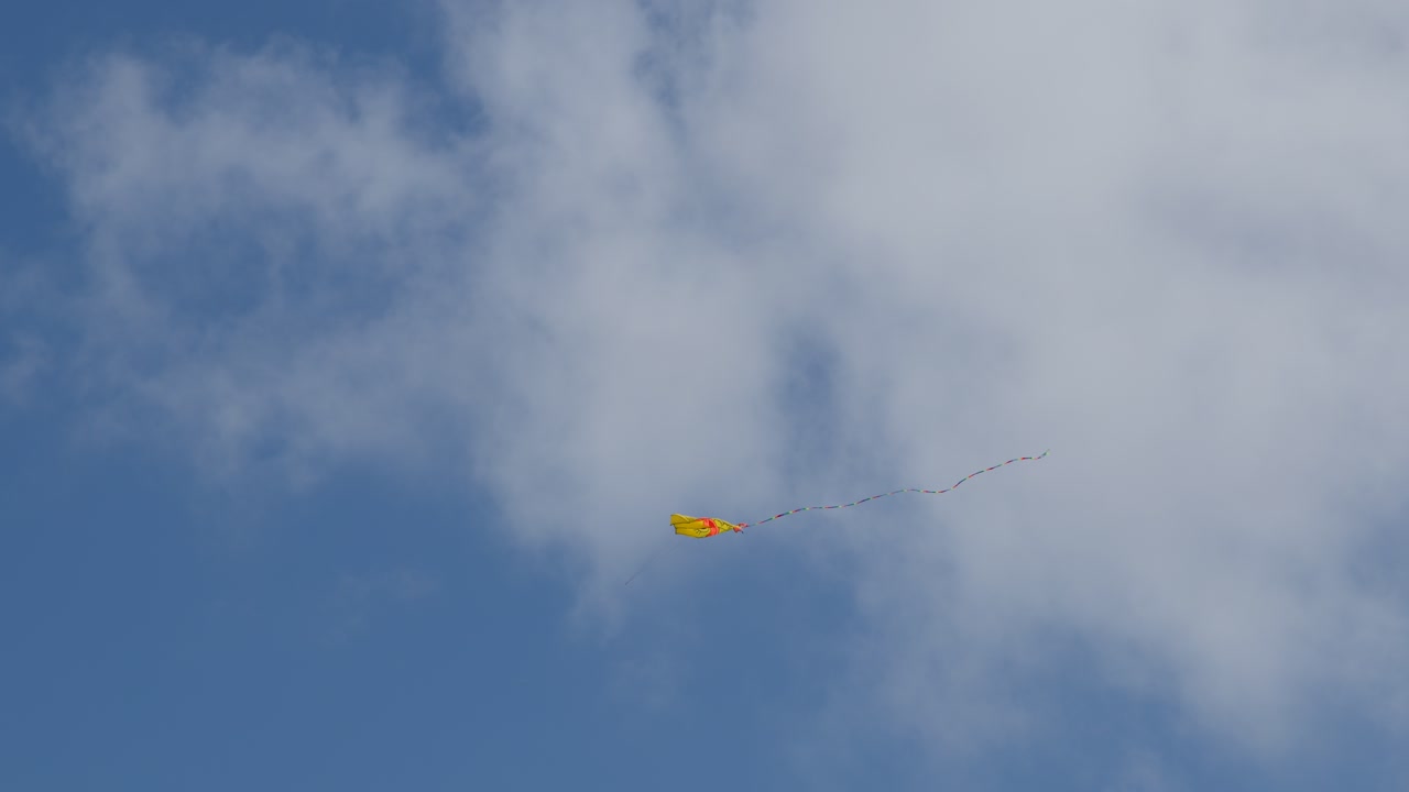 Yellow kite with colorful tail soars in daylight, drifting across blue sky and clouds