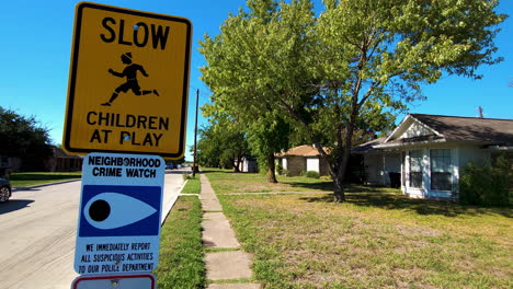 Leaves-rustle-on-trees-in-the-background-of-a-neighborhood-street,-a-street-sign-says-to-slow-down-for-children-along-with-a-crime-watch-reminder