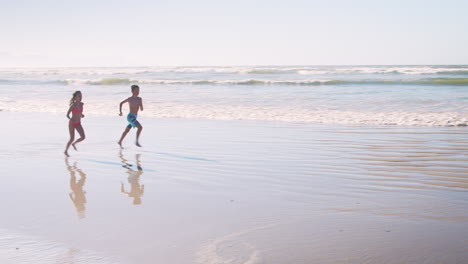 Niño-Y-Niña-En-Vacaciones-De-Verano-Corriendo-Juntos-Por-La-Playa-Hacia-La-Cámara