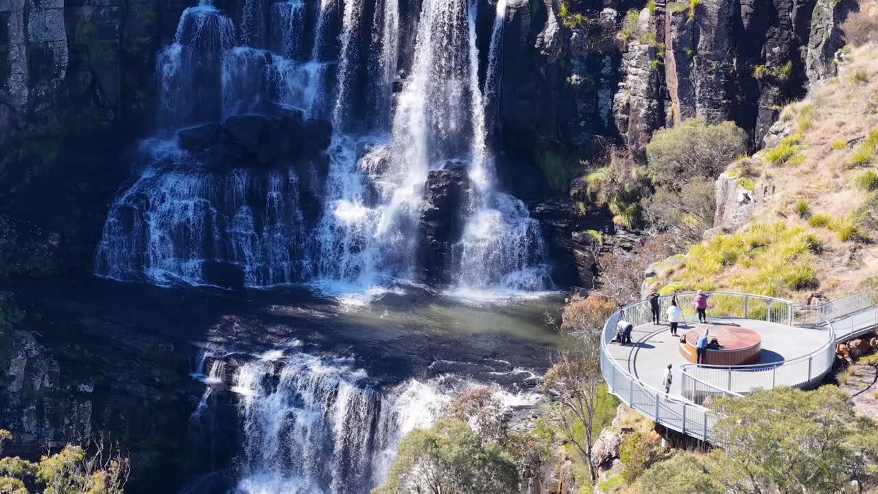 A group of people observe a multi-tiered waterfall from a modern clifftop lookout platform on a sunny day, surrounded by rugged natural landscape. Aerial perspective, steady camera
