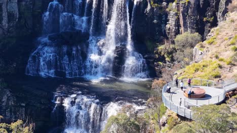 a group of people observe a multi-tiered waterfall from a modern clifftop lookout platform on a sunny day, surrounded by rugged natural landscape. aerial perspective, steady camera
