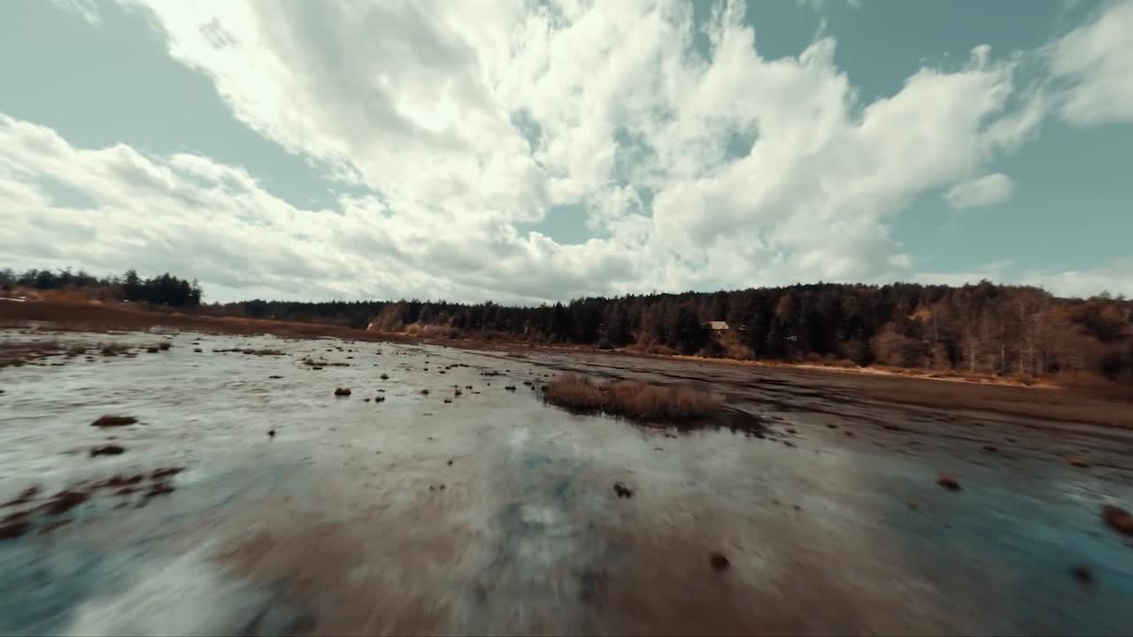 FPV aerial drone flying close to wetland or bog marshland reflective water during autumn cloudy day with trees, landscape and clouds reflecting on the water while drone flies forward and tilts.