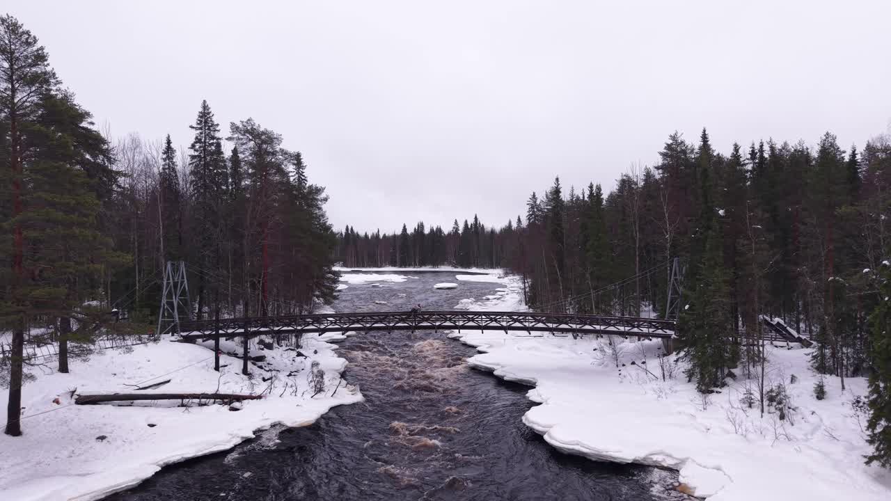 Una pareja caminando por un puente sobre un río con un tono marrón rojizo que fluye a través de un bosque de coníferas, Finlandia, vista aérea