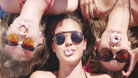 vista superior de tres amigas adolescentes acostadas de espaldas soplando burbujas de caramelo en la playa