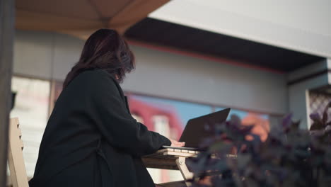 professional woman in black coat works on laptop outdoors in urban setting with decorative pillar and blurred cityscape in background