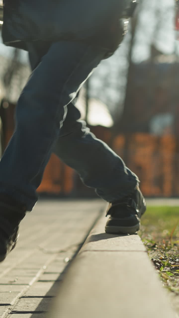 A close-up shot of a child walking along a pavement, holding a stick and missing a step while balancing, the child is dressed in a shiny black jacket