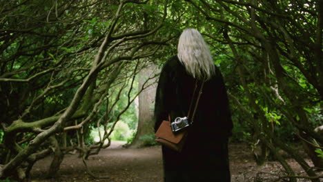 young blond photographer hipster girl woman walking through a magical story forest with scary trees with vintage bag and camera fujifilm old photographic camera capturing adventure european summer