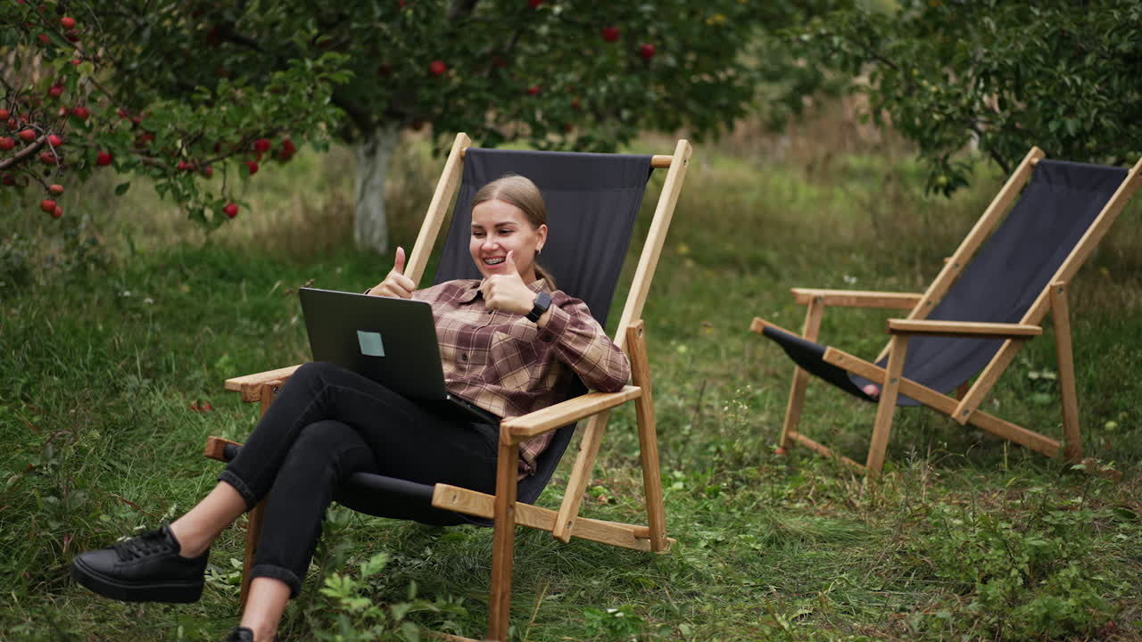 Cheerful lady having online video chat and is happy about something. Woman rejoices raising her hands and smiling happily. Nature at backdrop.