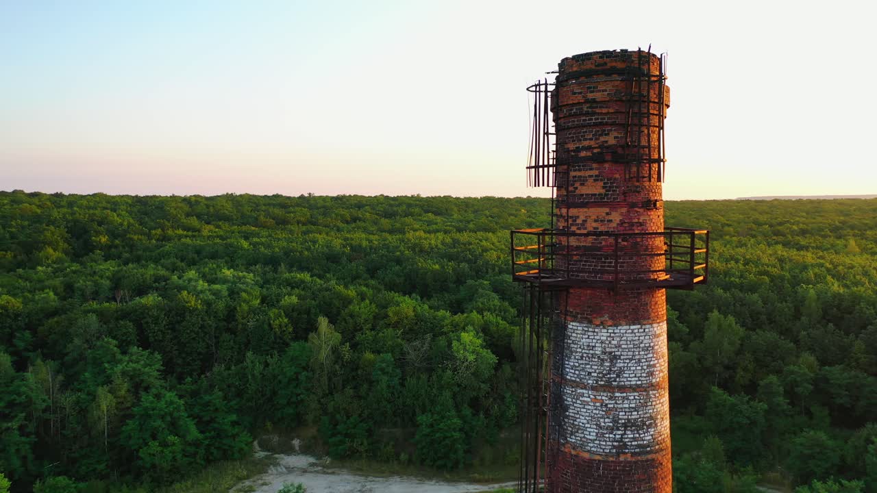 Brick industrial tower on nature background. Old pipe in ruined plant at sunset. Chimney surrounded by green trees and river in the countryside. Aerial view.