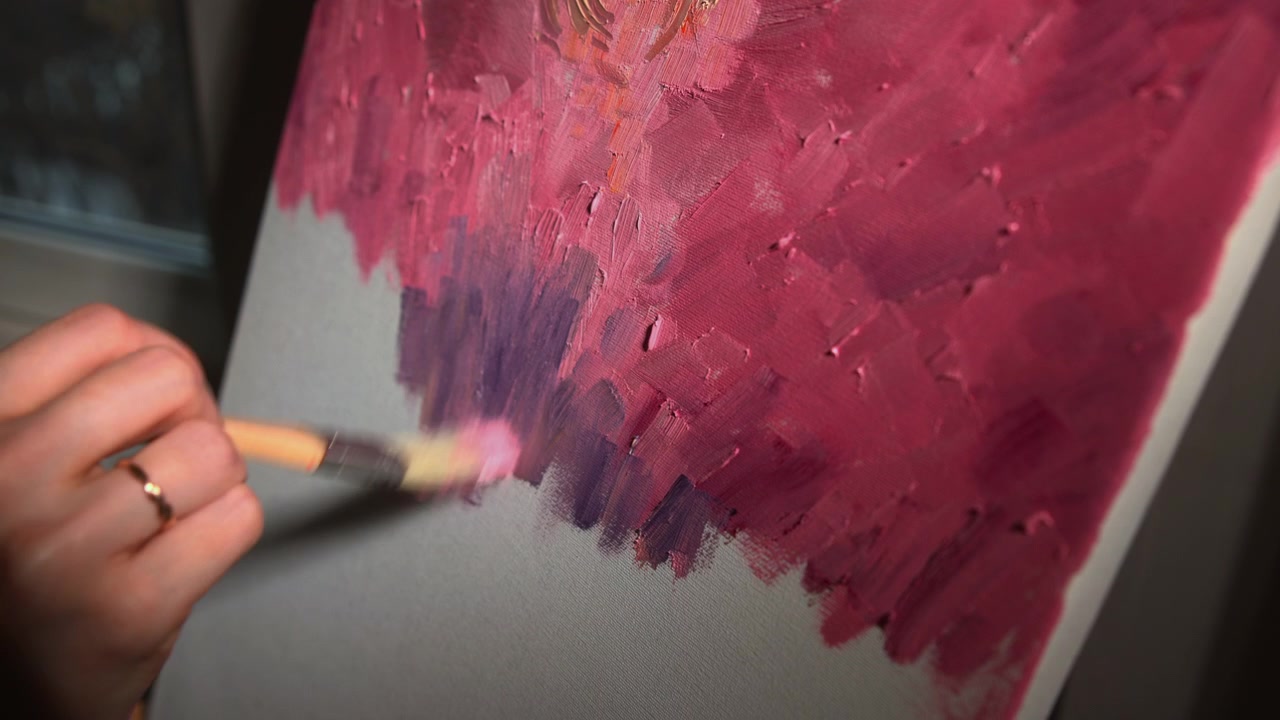 Primo piano di una mano che applica pittura spessa magenta e viola con un pennello su una tela, creando pennellate astratte a strati in un ambiente interno illuminato delicatamente.