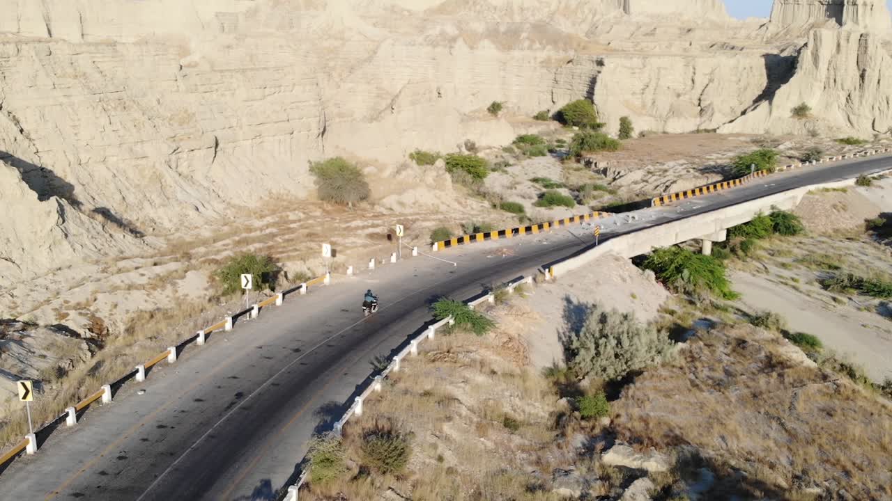발루치스탄 사막 풍경에 있는 힌골 국립공원을 통해 빈 곡선 고속도로를 따라 운전하는 오토바이 뒤에서 공중 추적 샷