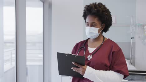 african american female doctor wearing face mask and holding documents in hospital room, slow motion