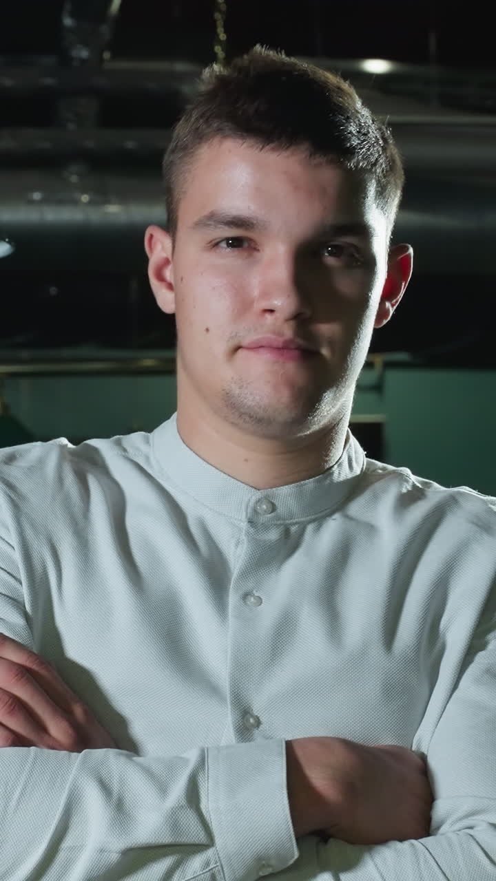 primer plano de un caballero con camisa blanca con los brazos cruzados y una sonrisa ligera en la sala de billar. múltiples mesas de billar verdes, lámparas aéreas y un interior moderno crean una atmósfera profesional pero relajada