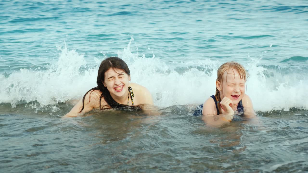 한 소녀와 함께 젊은 활발한 어머니는 해변에서 바다 파도에서 재미를 즐기고, 웃고, 모래놀이를 하고 있습니다.