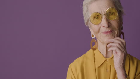 senior woman with short hair wearing mustard colored shirt and jacket, earrings and sunglasses posing with hand on chin and looking at camera on purple background