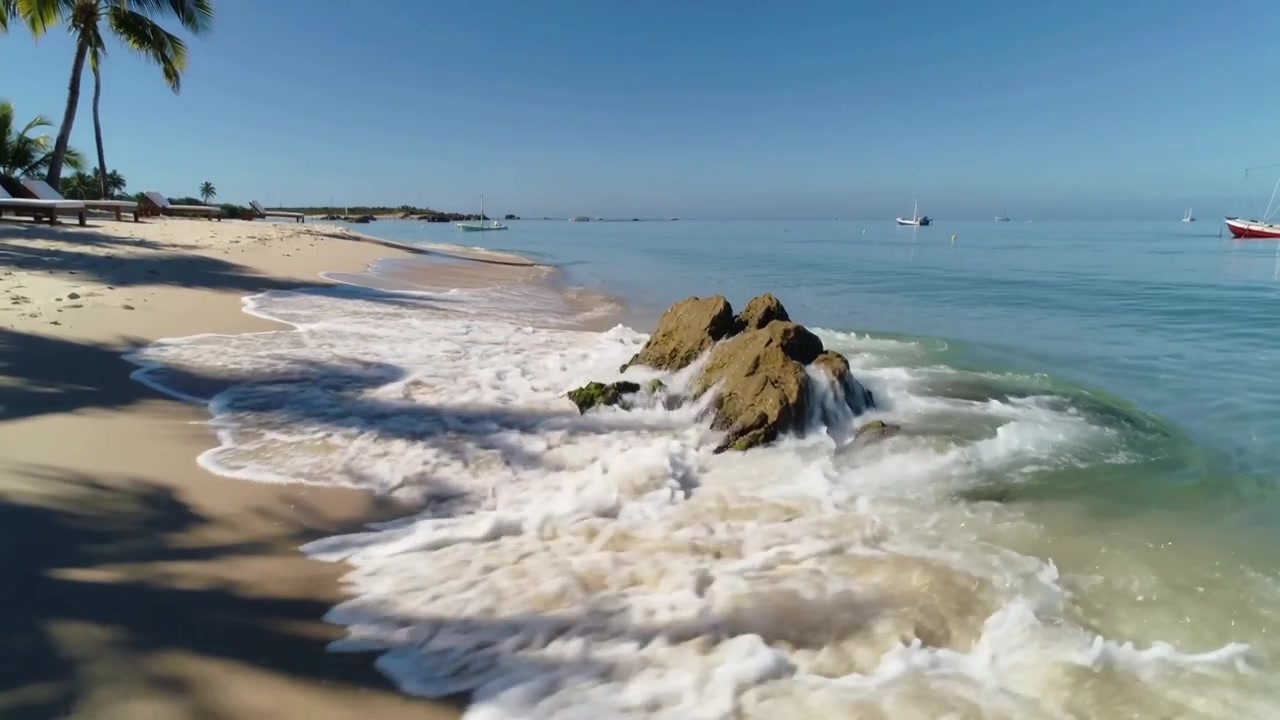 Onda forte quebrando em afloramento rochoso na costa, espalhando espuma branca sobre a areia, com espaço para texto.