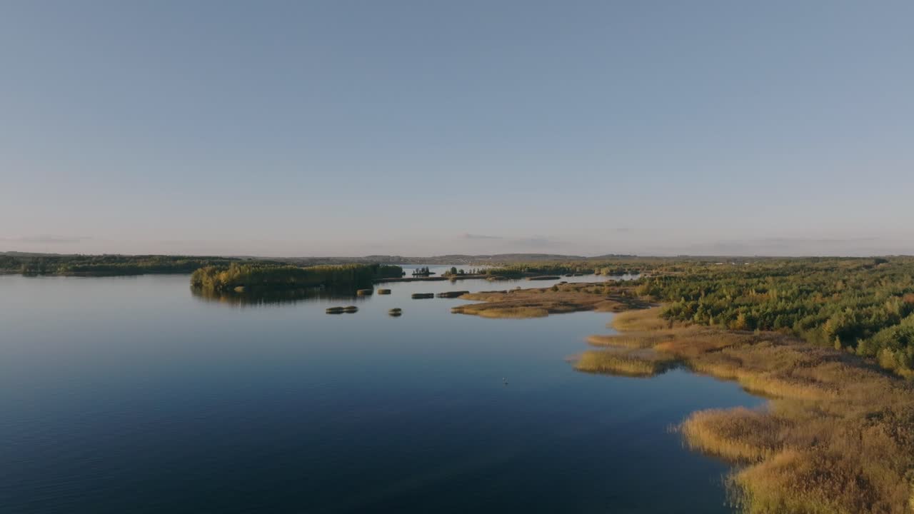 The drone shot shows a lake and its shoreline as the drone flies forward. The calm water and the coast come into view, with a clear, cloudless sky stretching above.