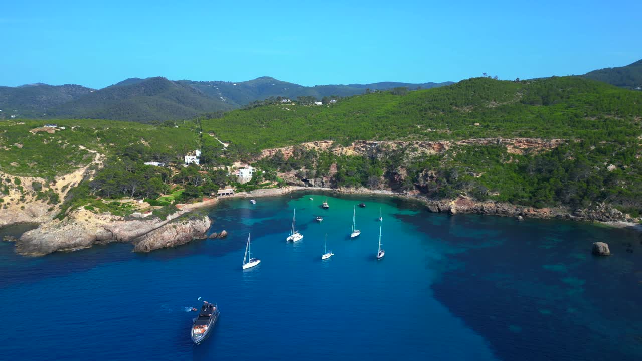 sailboats and yachts moored in turquoise waters of a bay on Ibiza, Spain, surrounded by lush greenery. Gorgeous aerial view flight panorama orbit drone