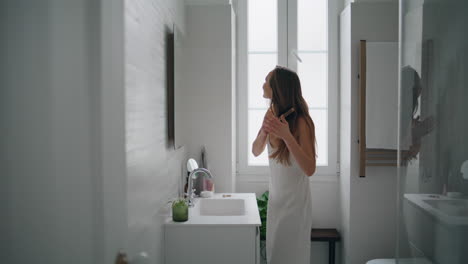 Young-woman-brushing-hair-in-bathroom.-Tender-girl-applying-mask-after-shower