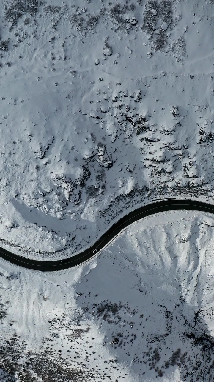 A winding road meanders through a snow-covered landscape. The aerial view captures the contrast between the dark road and the white snow. Suitable for travel and winter-themed projects.