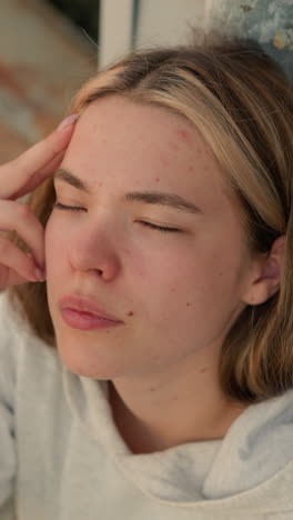 primer plano de una mujer joven descansando su mano por su cabeza, pareciendo perdida en profundos pensamientos, parpadea sus ojos, con una expresión tranquila e introspectiva, contra un fondo de pasos desgastados