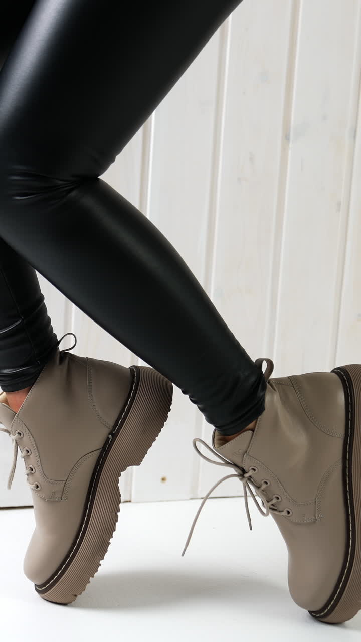 Demonstration of low beige boots with laces. Model in black leather pants jumps up in fashionable footwear. White wall backdrop. Vertical video
