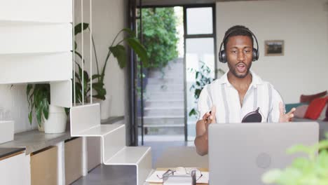 happy african american man sitting at table in kitchen, using laptop and making vlog