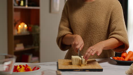 close up of woman at home in kitchen preparing healthy vegetarian or vegan meal slicing tofu on board with knife