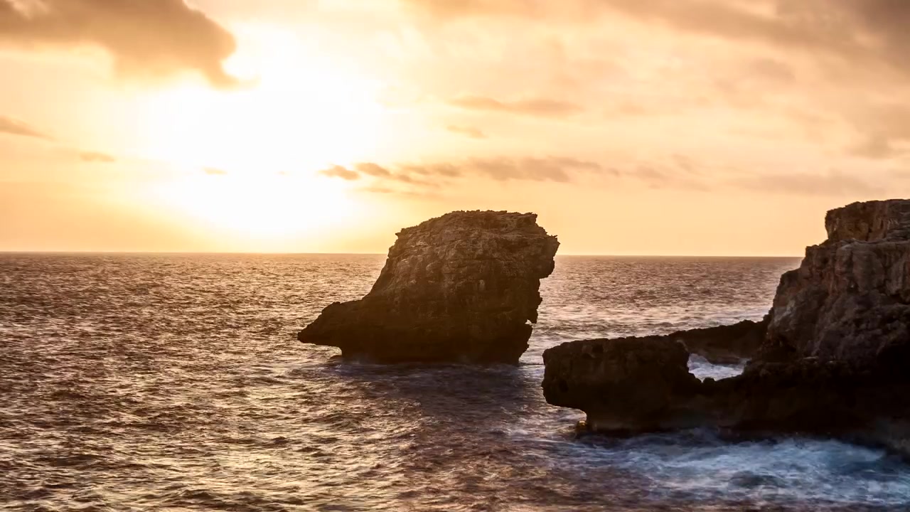 A água do oceano embate nas rochas ao pôr do sol em Binibeca, Menorca, com um brilho dourado de luz no céu e um raio a mergulhar na água