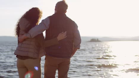 Vista-Trasera-De-Una-Pareja-Romántica-Abrazándose-Y-Viendo-La-Puesta-De-Sol-Desde-La-Orilla-Del-Lago