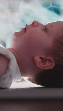 little boy lies down on floor against bright lamp ready to lift heavy barbell. motivated toddler enjoys workout with weight in gym with smoke slow motion