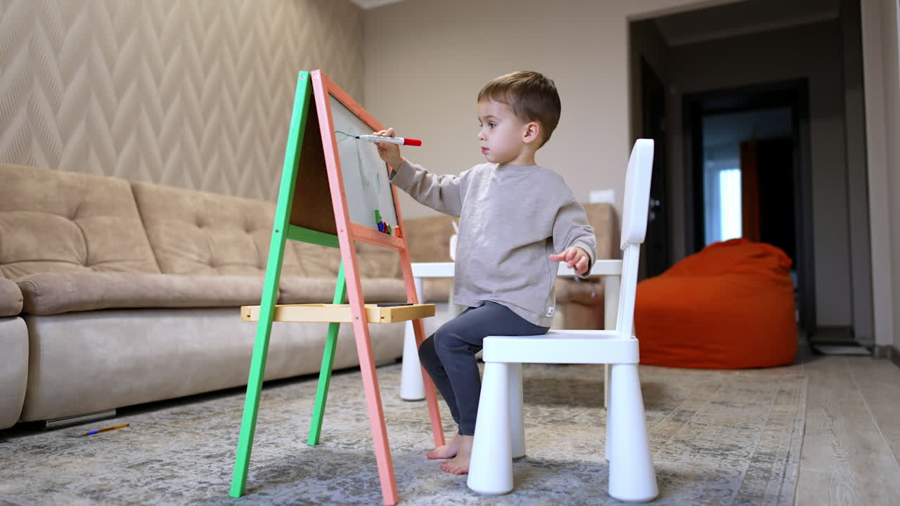Caucasian toddler sitting on the chair in front of the whiteboard. Cute kid drawing with a marker.