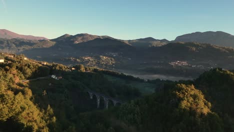 aerial-drone-view-of-a-bridge-in-a-forest-with-mist-at-golden-hour-in-morning,-hill-and-mountain-in-background,-tuscany,-italy,-europe