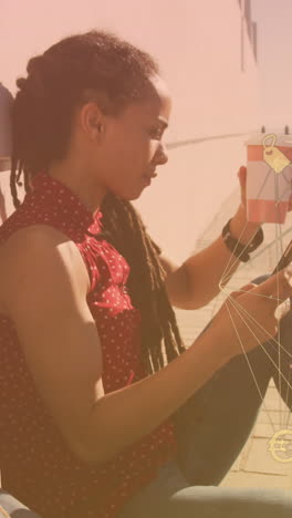 globe of digital icons against african american woman using smartphone sitting on the promenade