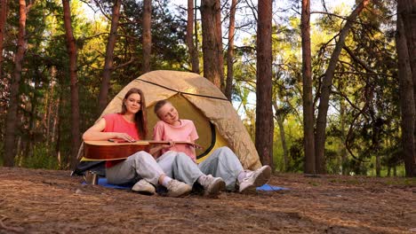 two girls playing guitar at a forest campsite