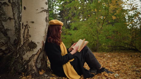 side view of stylishly dressed woman sitting alone, leaning against tree, flipping through pages of book in peaceful autumn setting, surrounded by golden leaves and tranquil forest atmosphere