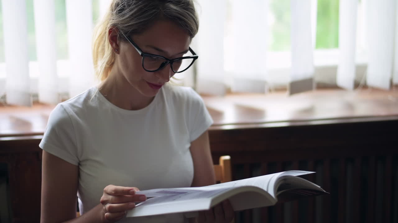 Blonde woman wearing black reading glasses and a white T-shirt, reading a book on a chair in front of a window