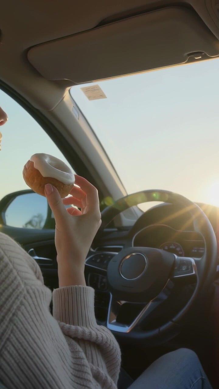 Video verticale: La fame che spinge una donna con un top beige a portare una ciambella glassata alle labbra nel bagliore della ruota dell'auto.
