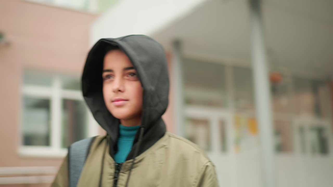 후드 재킷을 입은 젊은 학생이 캠퍼스 근처를 걷고 있는 측면 모습이에요. 흐릿하게 보이는 학술 건물 배경은 일상적인 학교 분위기와 젊은 라이프스타일을 반영하고 있어요.