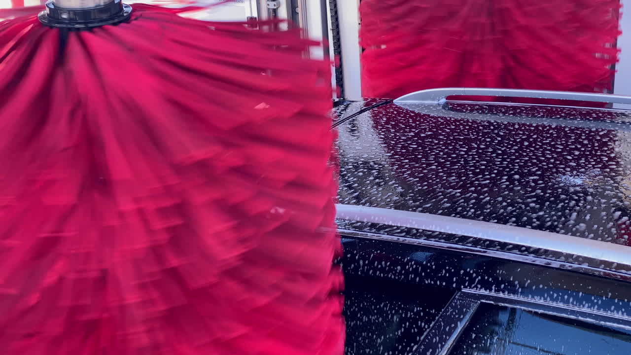 Tetto dell'auto coperto di schiuma di sapone e bolle mentre spazzole rosse rotanti puliscono il veicolo in una postazione di autolavaggio automatico.