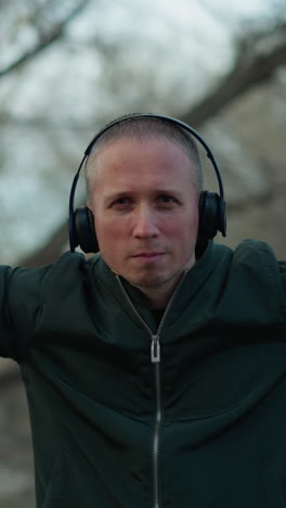 un hombre con una chaqueta verde y auriculares baila con movimientos expresivos de la mano alrededor de su cara en un bosque sereno