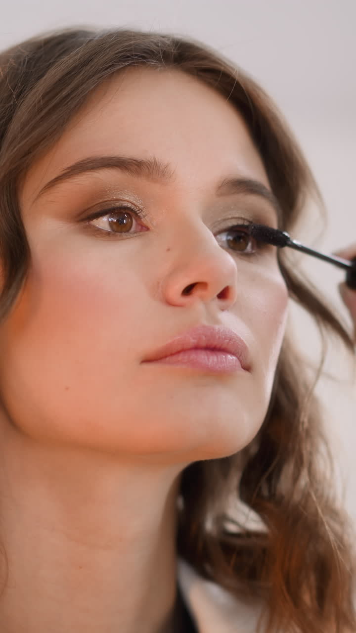 Focused woman applies black mascara on eyelashes with wand on blurred background. Redhead lady enjoys doing makeup for fashion event at home closeup